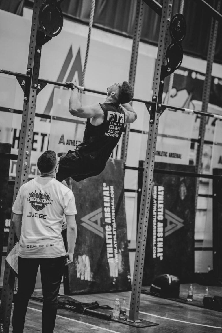 Athlete performing pull-ups in a competitive gym environment under judge's supervision.
